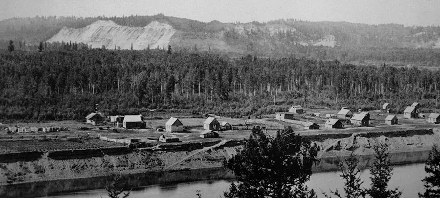 Black-and-white photo of a row of wooden houses on a river terrace, with forest and pale silt bluffs behind