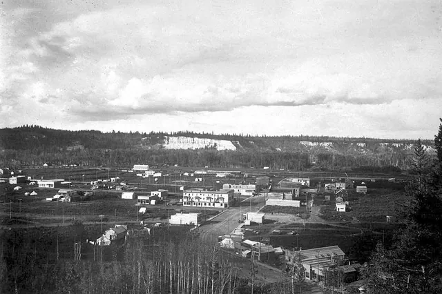 Historic black and white photograph of George Street in early Prince George, with wooden storefronts and a dirt road