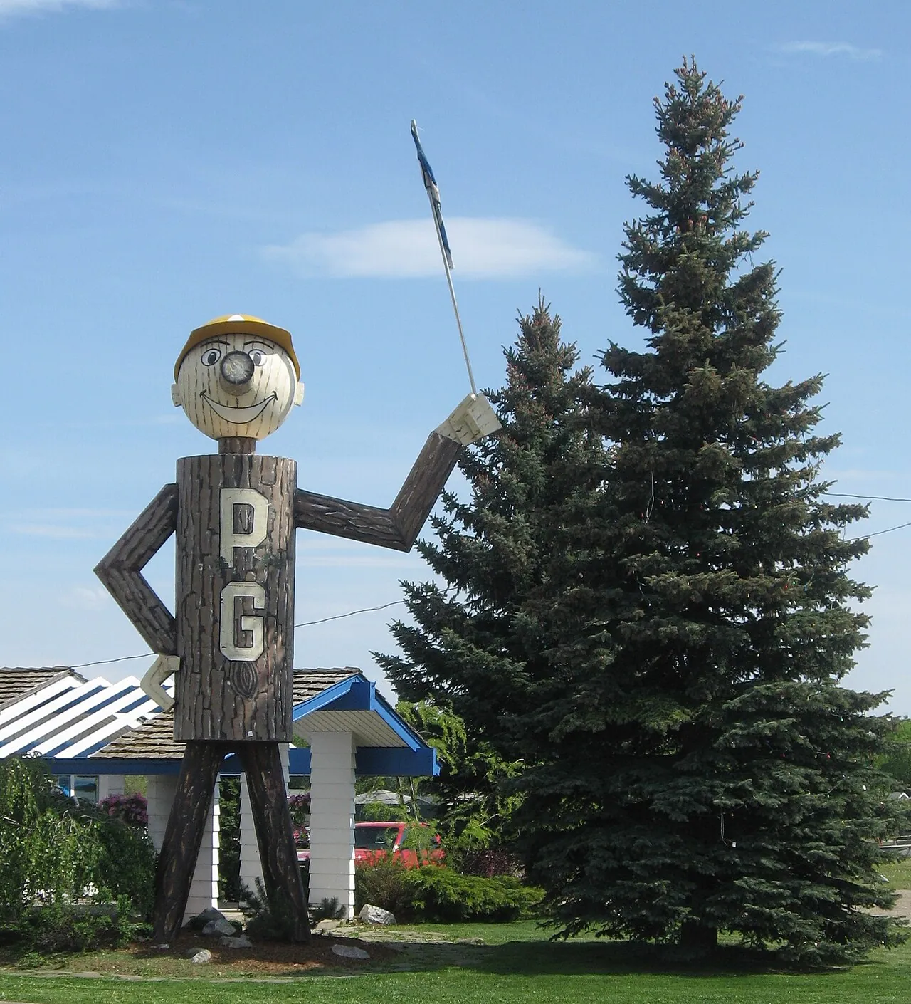 Mr. PG, the city’s tall lumberjack-style mascot with a round head and a PG logo, standing beside a spruce tree in Prince George