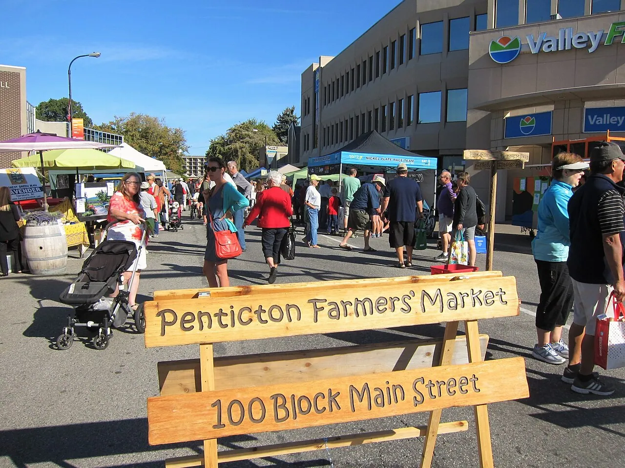 Shoppers browsing stalls at a busy outdoor farmers’ market (Penticton, shown as a generic example of a BC market)